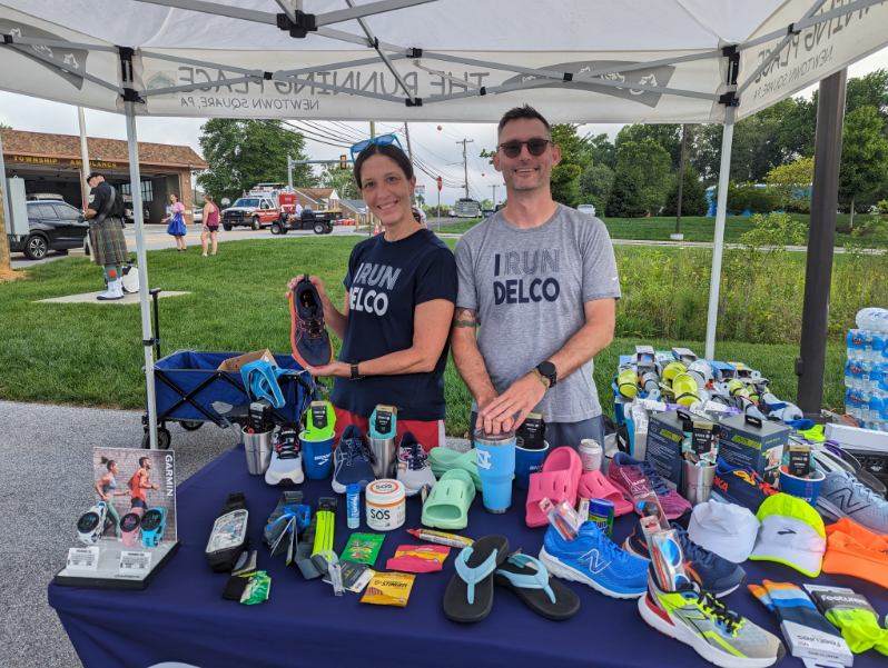 Photo of TRP owner Ken Taylor and Shoe Seller Ann under a tent at a race. Ann is showing off a shoe and there is a table of shoes and socks spread out in front of them.